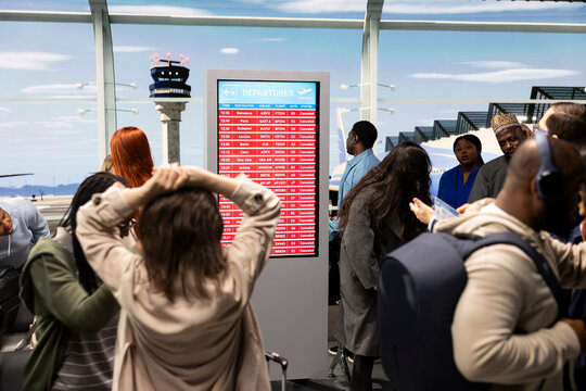 Passengers in airport worried about disruption causing delayed flights, reacting to red alert on airline screen. International travelers express anxiety after flight cancellation, receiving bad news