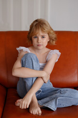 Fototapeta premium Young girl, fair skin, blonde hair, casual clothes, sitting with legs crossed on orange leather sofa, front view, look at camera, bright room. Concept of childhood, home life, calm mood.