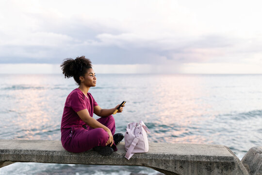 Young Woman Sitting by the Water in Scrubs and Using Her Phone
