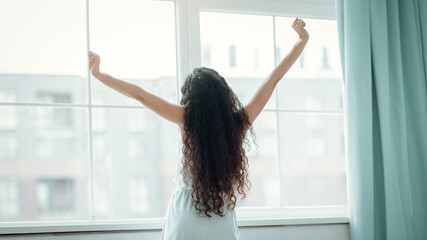 A woman stands by the window with her arms raised. She has long hair and is enjoying a moment of stretching as sunlight comes through the window in a city apartment. © Prostock-studio