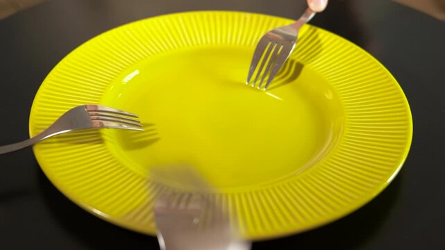 Many forks knocking on an empty yellow plate. Multiple forks held by different hands simultaneously knock on an empty yellow plate on a dark table, symbolizing hunger, waiting for food, impatience