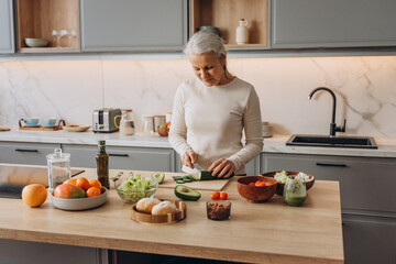 Happy elderly housewife cuts fresh vegetables for tasty salad at wooden countertop