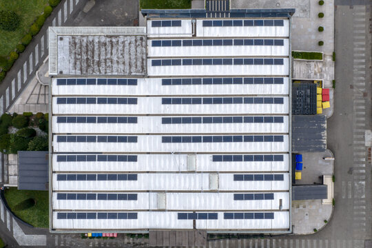 Aerial view of rows of solar panels installed on a white industrial building roof with surrounding paved areas and greenery in Reggello, Tuscany, Italy.