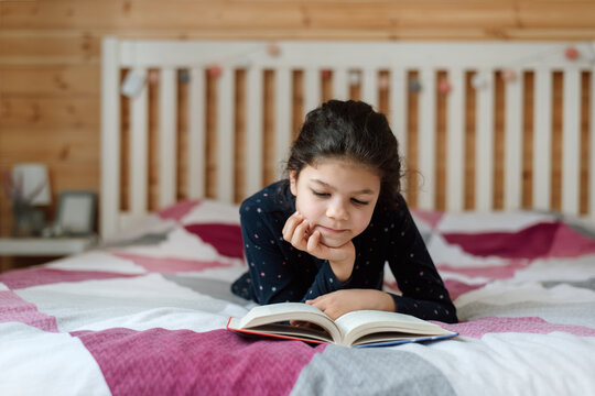 Portrait of a girl reading a book in her bedroom lying on her bed.