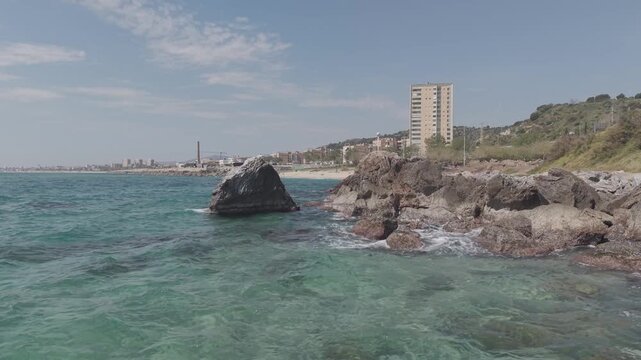 30 seconds aerial drone video of Montgat beach shoreline with gentle Mediterranean waves, sandy coast and seaside buildings, Playa de las Rocas in El Maresme, Spain under clear blue sky. Long shot.