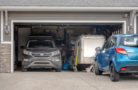 Calgary, AB, Canada. April 15, 2026. A packed suburban garage showing a Honda CRV, cargo trailer, and blue Mazda 2 parked on the driveway.