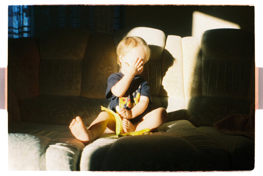 Toddler Sitting on Sofa in Warm Sunlight Closes Face With Hand
