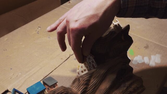 A close-up shot of hands carefully arranging miniature wooden houses and a windmill on a beautifully textured, dark wood driftwood base.