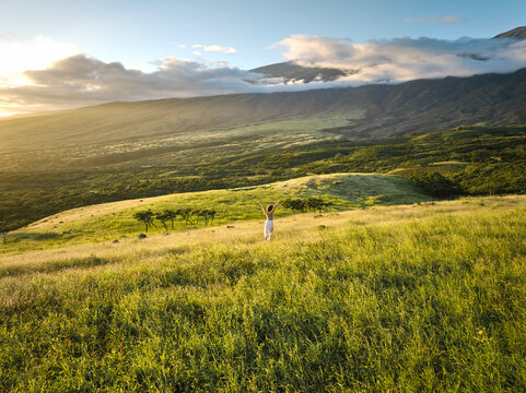 Woman in White Dress Walking Through Lush Green Grass Valley at Sunset