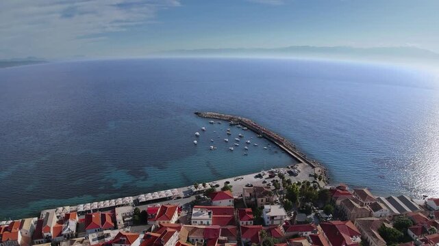 Aerial drone view of small harbor with breakwater and anchored boats in clear blue Mediterranean sea, coastal town in Greece, sunny summer day, travel destination