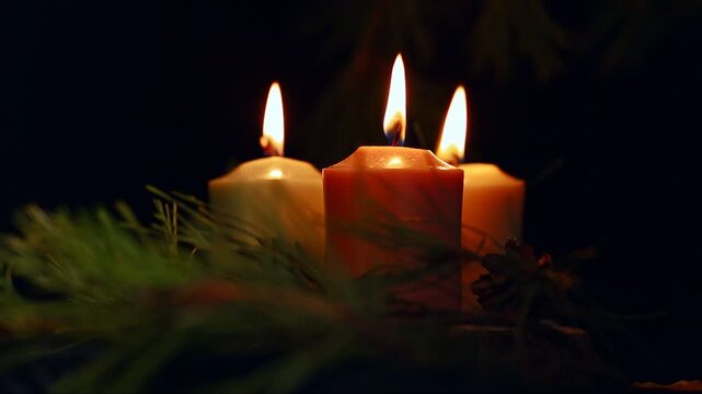 Three colorful Christmas candle flames - pink, green, yellow burning near green fir branch on wood candlestick with dried orange, cones on black background. Happy New Year celebration. No people. 