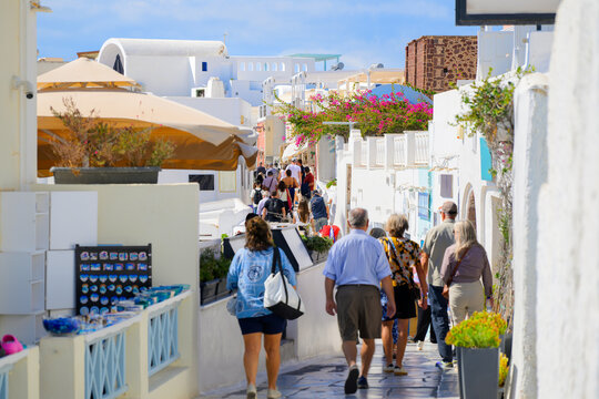 A crowded narrow pedestrian walkway past whitewashed buildings with shops and cafes in the picturesque center of Oia, Greece, on the island of Santorini.