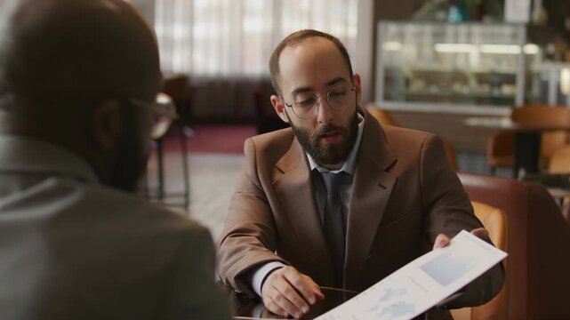 Caucasian businessman and his African American colleague shaking hands and discussing financial report with data charts during meeting in hotel lounge