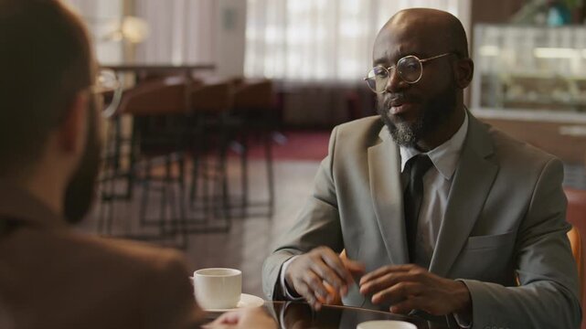 African American businessman in formalwear and glasses discussing agreement and shaking hands with colleague after sealing deal during meeting in restaurant. Over the shoulder shot