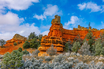Striking tall sandstone hoodoo and layered red cliffs in Red Canyon, Utah. Vibrant orange rock contrasts with green and snow-dusted vegetation under blue sky. © ZL Visuals
