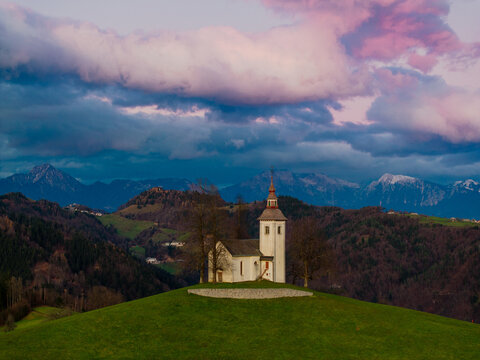 Aerial view of Saint Thomas Church on a green hilltop with the Julian Alps and colorful sunset clouds in the background Slovenia.