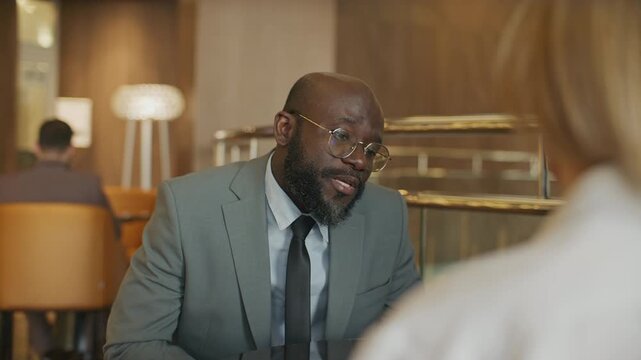 Black business executive in suit and glasses talking to colleague during informal meeting in modern hotel lounge, explaining project key points and sharing ideas. Over the shoulder view
