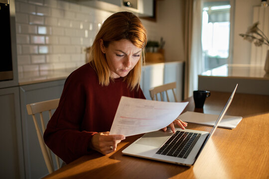 Focused adult reviewing bills at laptop in home kitchen