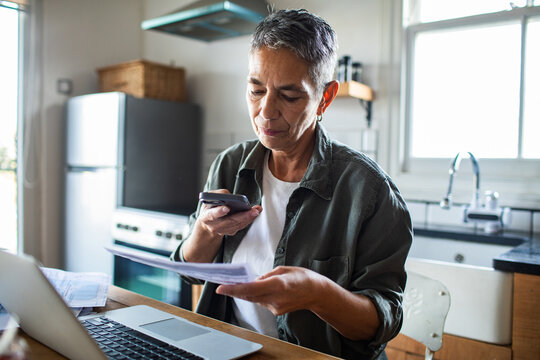 Mature woman managing household finances by phone in home kitchen