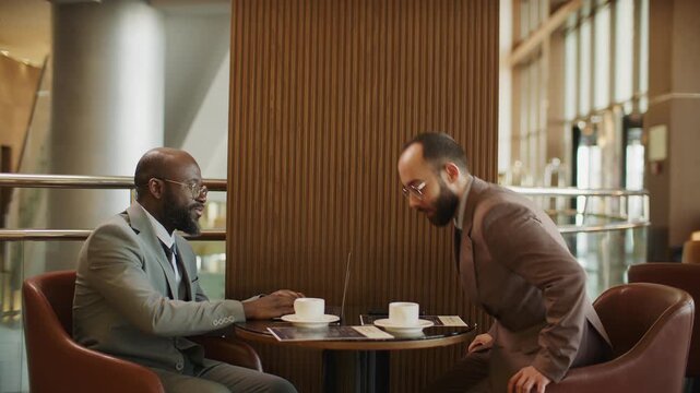 African American business advisor meeting with client in hotel lounge, greeting him with handshake, using laptop and providing consultation at table with coffee cups and papers