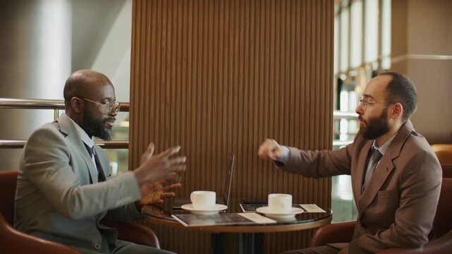 Two diverse business partners discussing contract over coffee and sealing deal with handshake during meeting in cafe or hotel lounge