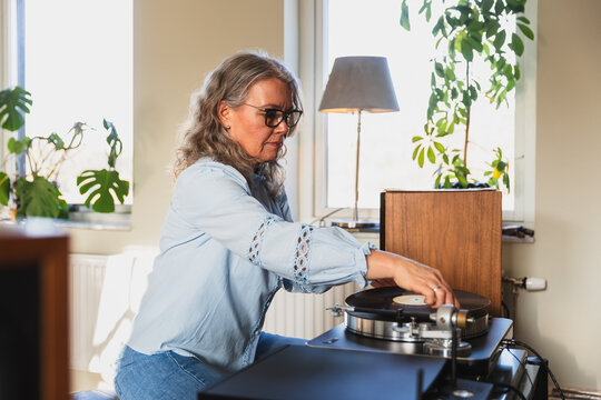 Mature woman playing vinyl record on turntable in bright living room