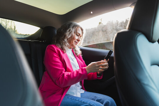 Professional woman in pink blazer using smartphone in back car seat 