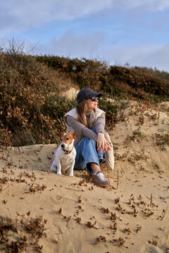 Young Woman Sitting on Sand Dune With Her Dog Relaxing on beach.