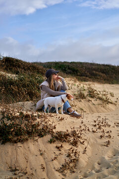 Young Woman Sitting on Sand Dune With Her Dog Relaxing in Casual Autum