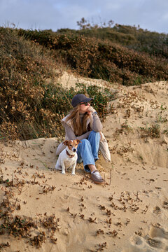 Young Woman Sitting on Sand Dune With Her Dog Relaxing in Casual Autum