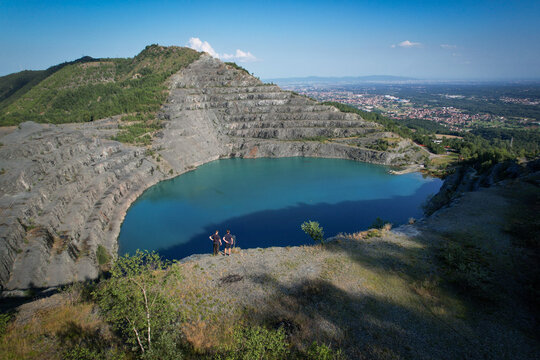 Aerial view of the Amiantifera di Balangero asbestos mine featuring a turquoise lake, terraced rock walls, and two people standing on the edge in Balangero, Piedmont, Italy.