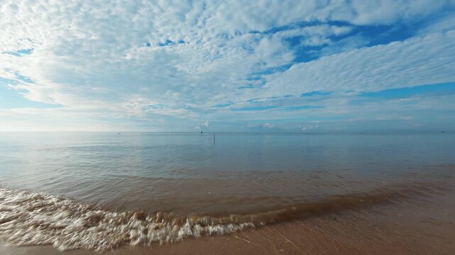 A wide landscape view of Seraya Beach in the morning, featuring the long shoreline, gentle waves lapping onto the wet sand, and the extensive blue sky covered with a textured layer of white clouds. Th