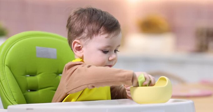 Cute little boy eating baby food on feeding chair at home. Camera moving right