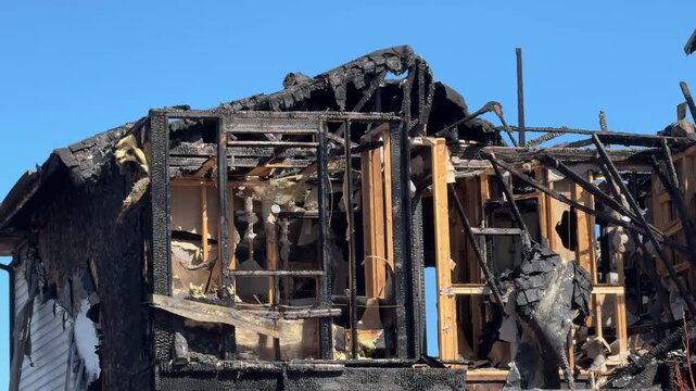 Aftermath of a house fire showing a charred, skeletal residential structure under a clear blue sky.