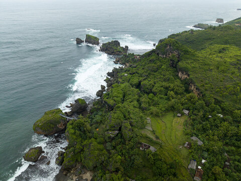 Aerial view of Siung Beach featuring rugged limestone cliffs, lush green vegetation, and white waves crashing against the rocky coastline in Tepus, Special Region of Yogyakarta, Indonesia.