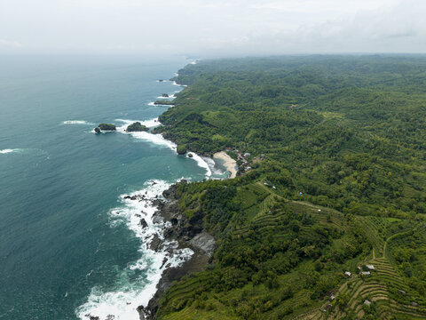 Aerial view of Siung Beach featuring a rugged coastline with turquoise ocean waves crashing against rocks, lush green hills, and terraced fields in Tepus, Special Region of Yogyakarta, Indonesia.
