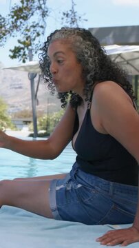 Vertical video: Calling woman in black swimsuit, denim shorts reaching pool showing yellow object