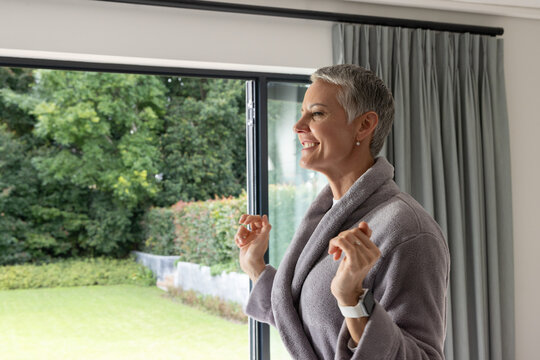 Senior female standing, looking out sliding glass door at home, gray robe, white watch, copy space