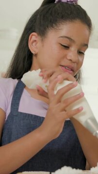 Vertical video: Twisting piping bag, teen in apron piping frosting on cakes at counter with mom