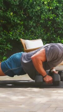 Vertical video: Starting man in shorts doing pushups and rows on patio with dumbbells for strength