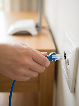 Close up of hand plugging blue ethernet cable into wall socket with router in background copy space