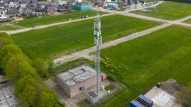 Aerial view of a tall telecommunications tower and utility building surrounded by green fields and a modern residential neighborhood in Almere, Flevoland, Netherlands.