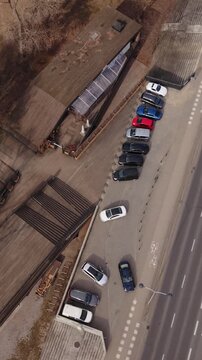 Vertical screen Aerial View Parked Cars Warszawa Street, Drone Overhead Captures Parallel Vehicles Along Curb, Triangular Wooden Structure And Muted Winter Trees, Quiet Traffic On Adjacent