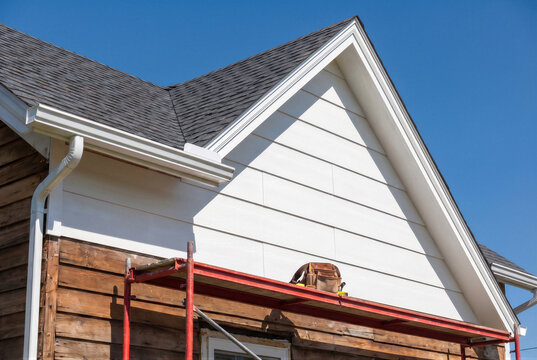 Exterior home renovation featuring new white horizontal siding installation on a gable end with scaffolding and a tool belt in place