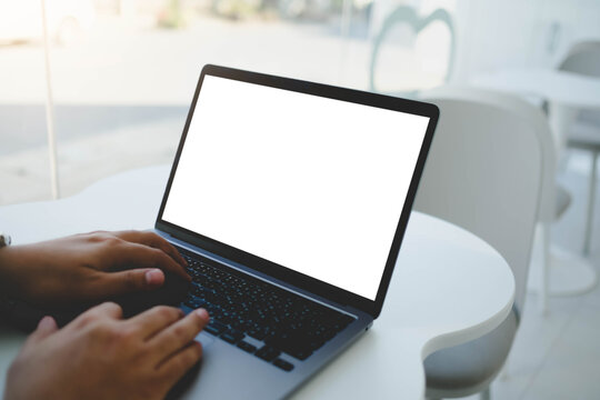 An over the shoulder view of a person typing on a laptop with a blank white mockup screen while sitting at a table in a bright indoor setting.
