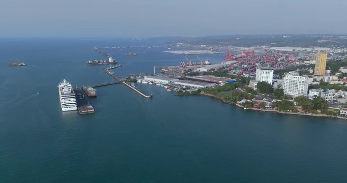 backward high angle aerial view of Sihanoukville autonomous port featuring a docked cruise ship, busy container terminals, and a cargo ship departing in the background.