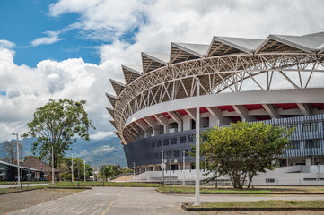 Modern National Stadium in La Sabana Park, San Jose, Costa Rica, iconic sports venue with steel architecture, surrounded by trees, city streets, and mountains under bright cloudy sky (February 2026). © J Duquette