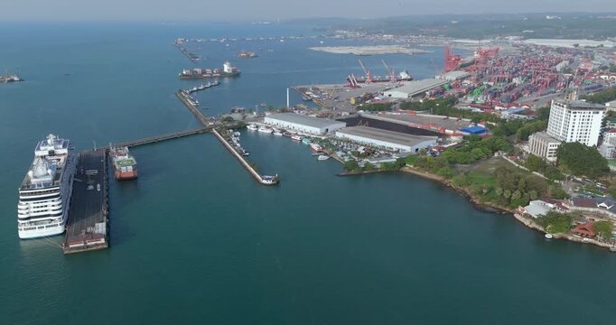 backward aerial view of Sihanoukville autonomous port featuring a docked cruise ship, busy container terminals, and a cargo ship departing in the background.