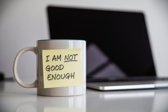 A white coffee mug with a yellow sticky note saying I am not good enough placed on a desk next to a blurred laptop representing imposter syndrome