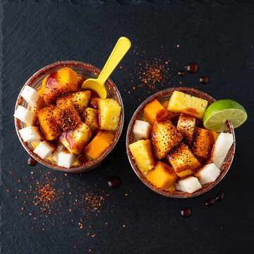 Mexican fruit cups (fruta con chili) with mango, pineapple, jicama, chamoy, and tajin on black background. Colorful snack concept.